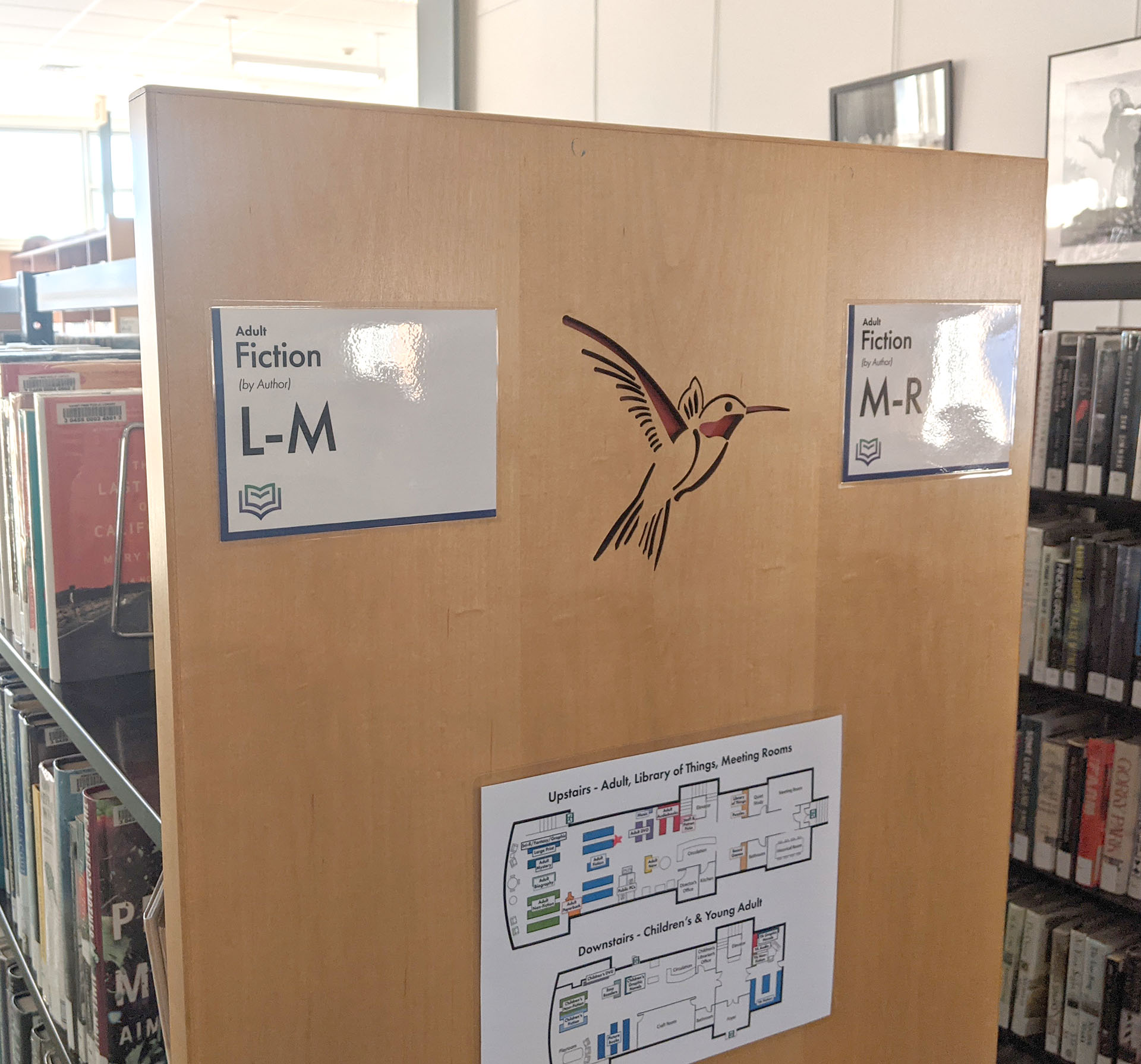 Signs at the end of a bookshelf that read
Fiction L-M and Fiction M-R, as well as a map of
the building below. There is a hummingbird carved
into the end of the wooden shelf